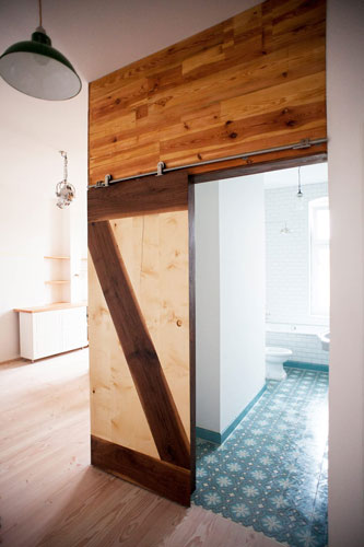 Open solid wood slide door on metal rail. View to bathroom with azure blue floor tiling. Wooden slide door with dark and light wood design pattern.