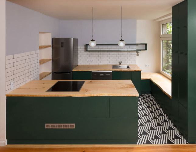 Complete kitchen with kitchen island in center. Worktop with sink and dishwasher, next to fridge. Wooden worktops, wooden lower cabinets painted green. Black and white patterned tile floor. front shot.