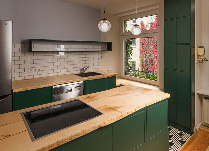 Complete kitchen with kitchen island in center. Worktop with sink and dishwasher, next to fridge. Wooden worktops, wooden lower cabinets painted green. Black and white patterned tile floor. Detailed view on storage wall cabinets.